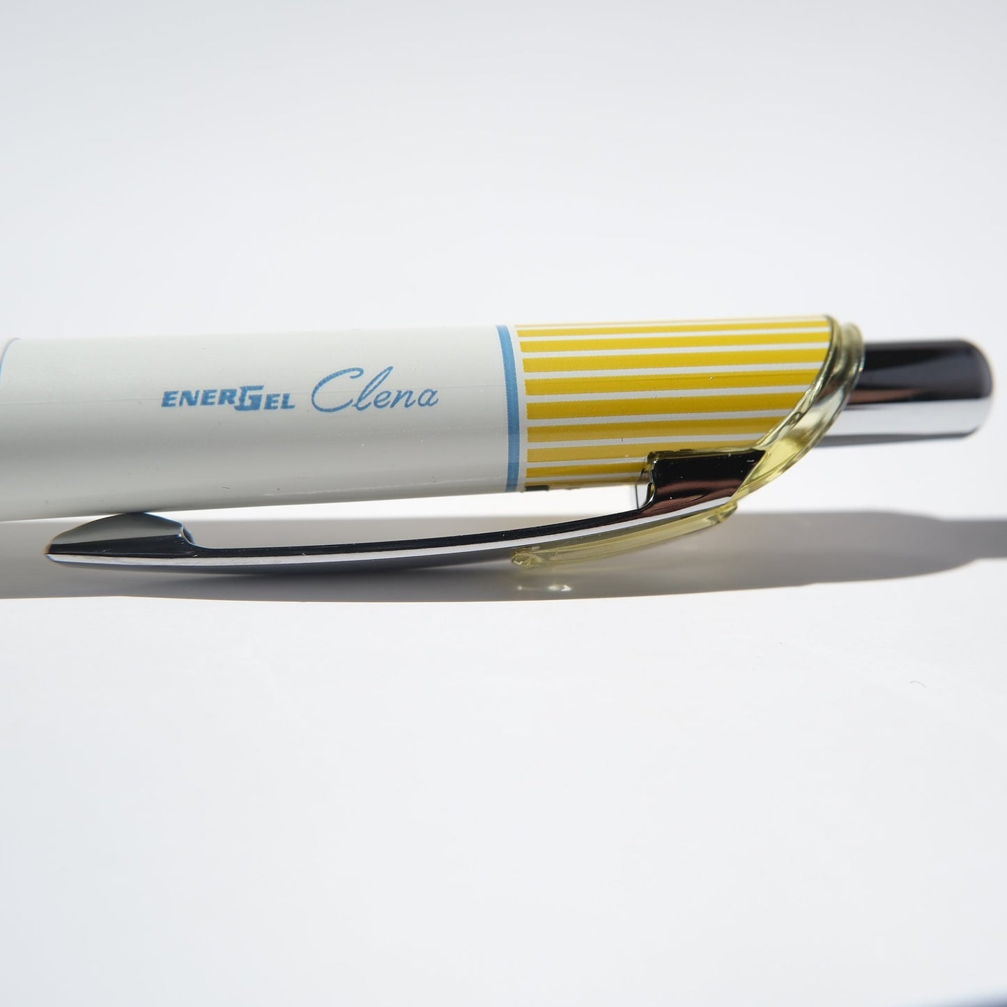 A close-up of a white EnerGel Clona retractable pen with a yellow striped clip and a silver tip, resting on a white surface.