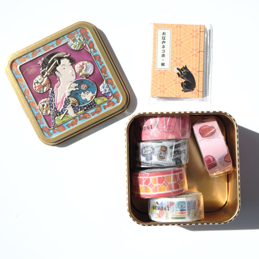 A styled flat-lay of a decorative metal tin set containing washi tapes. The set includes a closed tin with a Japanese geisha illustration on the lid, an orange card with a black cat, and an open tin showing four rolls of washi tape with pink, orange, and gray patterns.