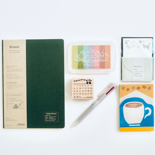 A flat-lay arrangement of stationery items on a white background, including a dark green and beige notebook, a small multicolor ink pad, a white envelope, a mini calendar, a pen, and a coffee-themed notepad.